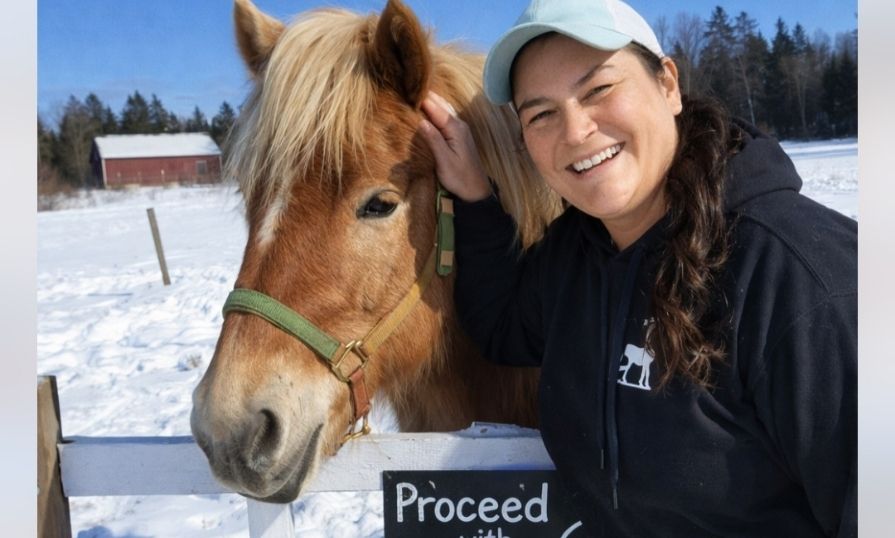 An Indigenous woman outside standing with a horse. The ground is snow covered and there's a red barn in the background.