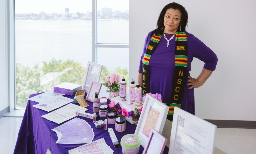 An African Canadian woman smiles as she stands behind a display of her natural products in decorative jars on a table covered with a purple tablecloth.