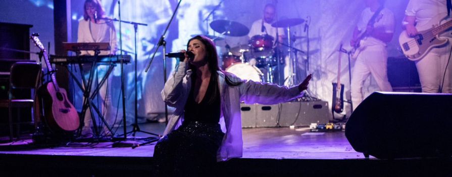 A concert stage with the female singer sitting on the edge of the stage singing and musicians in the background.