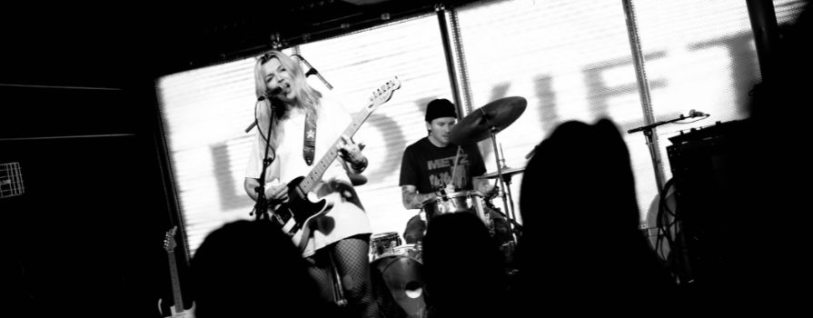 Photo from the crowd at a concert with a woman singing and playing guitar and man playing drums.