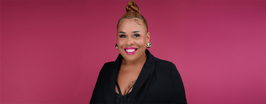 Tia Upshaw's headshot. Image is of a Black woman (Tia), wearing her hair in a bun and a black blazer. She's smiling at the camera in front of a deep pink background.