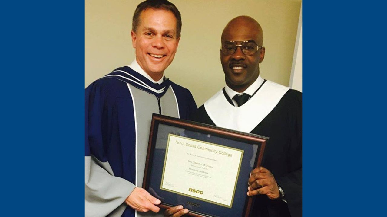 Two men standing together smiling at a graduation. One is holding a framed diploma.