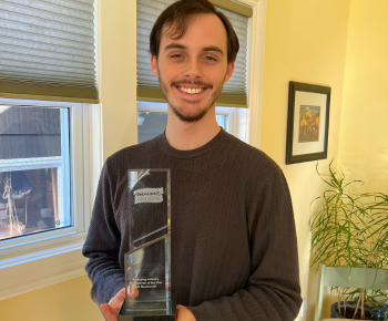 Nik MacDonald holding his award smiling in a living room.