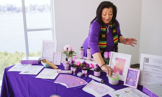An African Canadian woman smiles as she works on a display of products in decorative jars on a table covered with a purple tablecloth.