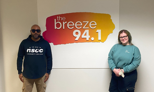 Male co-worker and Emma Freckelton standing on either side of The Breeze 94.1 sign on a wall.