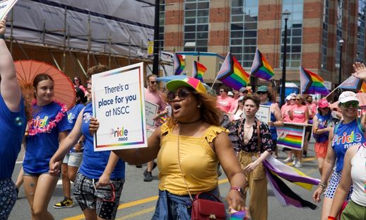 People march in a Pride parade holding rainbow flags and a sign that reads “There’s a place for you at NSCC.”