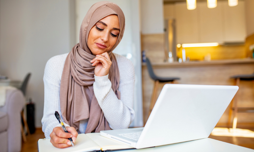 Woman wearing a hijab taking notes from a laptop screen.