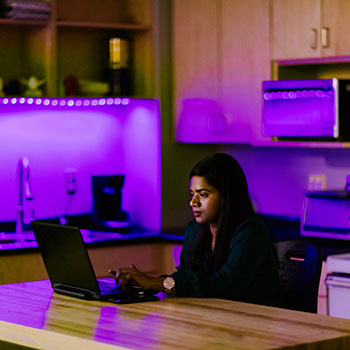 A young woman working on her computer in a kitchen area with colourful lighting.