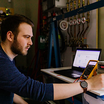 Male researcher surrounded by technology kneeling down in front of a laptop turning the dial on a piece of equipment.