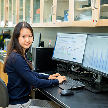 Female research assistant smiling slightly as she analyzing data on two computer monitors.