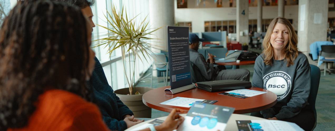 A woman in a Student Research Network shirt sits at a table in library smiling while she speaks with students