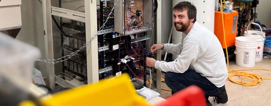 Male researcher kneeling in front of an electric vehicle battery system surrounded by other pieces of technology in a lab.