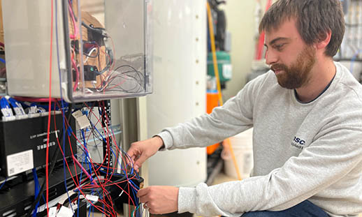 Male researcher kneeling in front of an EV battery system adjusting a collection of red and blue wires.