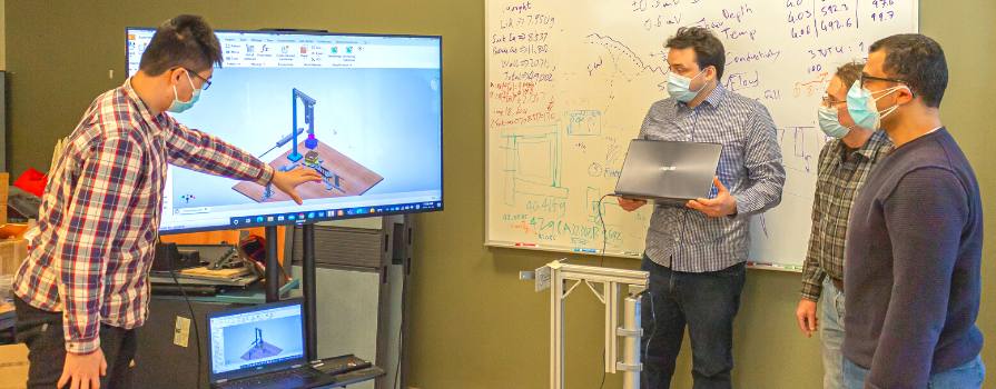Four men wearing medical masks are looking and discussing CAD design software on a large monitor. One man in a plaid shirt is gesturing towards the screen.