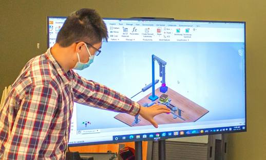 A man in a plaid shirt pointing at CAD design software on a large monitor.
