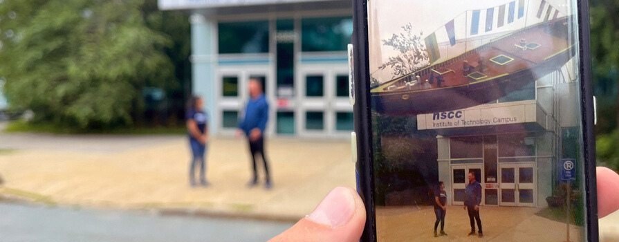 Person holds up smartphone by building entrance where a man and woman are chatting; screen shows rooftop pirate ship on the roof.