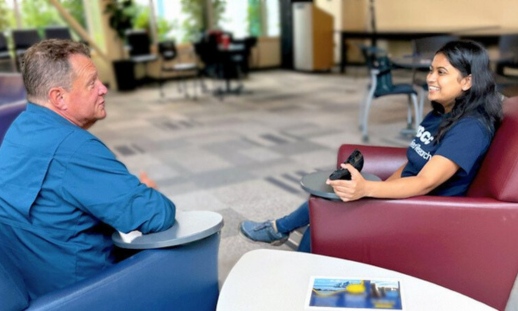 A man and woman sit in arm chairs in a college lobby engaged in conversation.