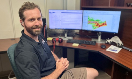 Man sitting at a computer with mapping software open, a lacrosse stick visible nearby.