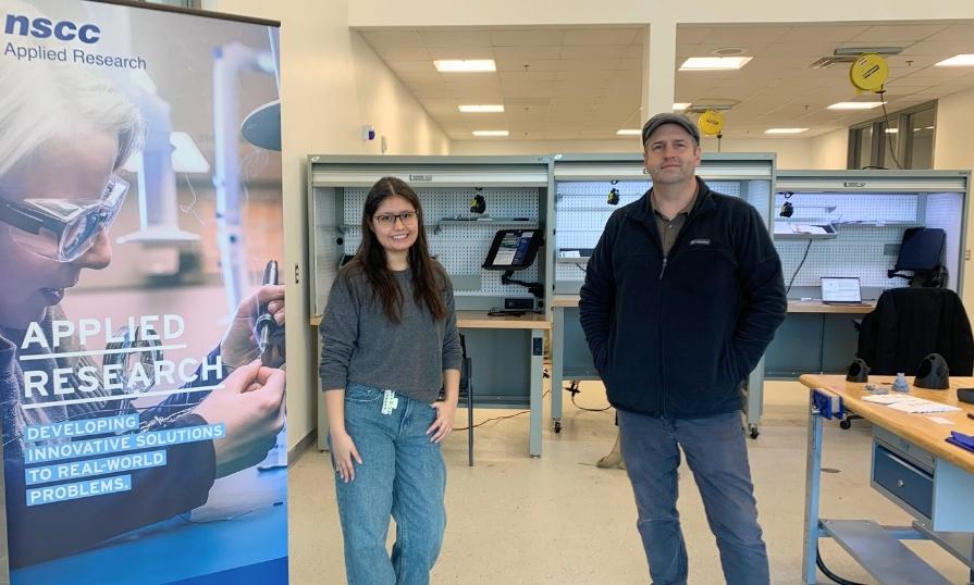 Two people standing in a lab space, smiling next to an Applied Research pull-up banner.