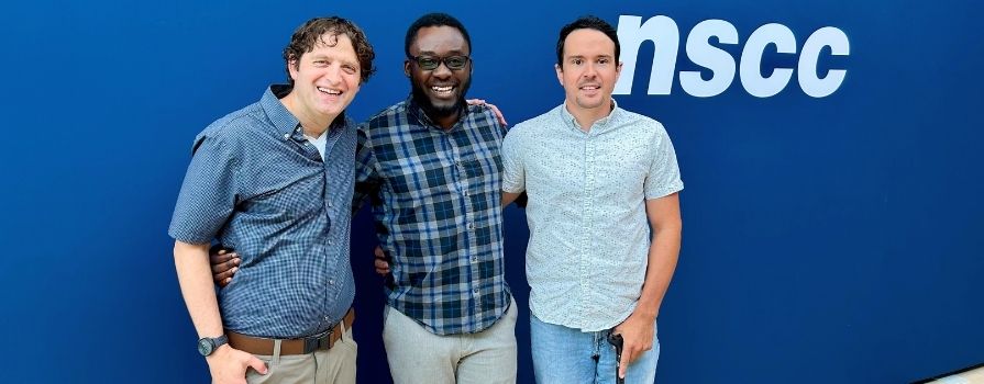 Three men with their arms around one another smiling in front of an NSCC banner.