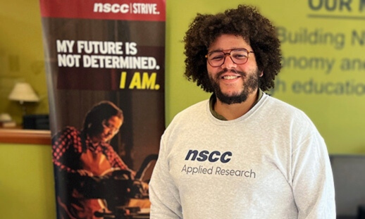 A young man, proudly smiling wearing glasses and an NSCC Applied Research branded sweatshirt. 