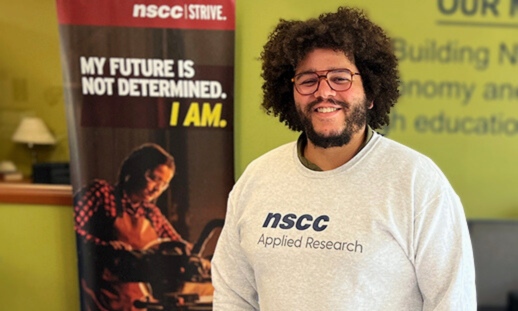 A young man, proudly smiling wearing glasses and an NSCC Applied Research branded sweatshirt.