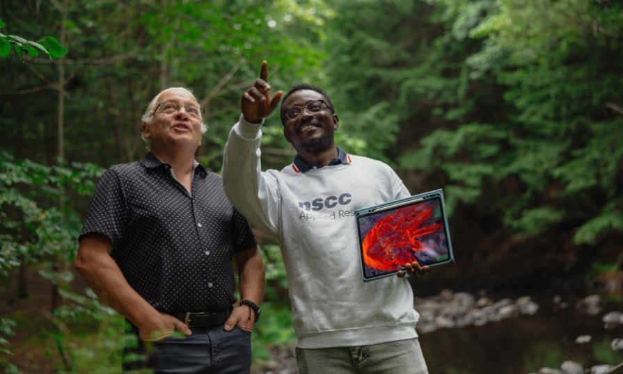 Two people stand in a wooded area, looking at a tablet displaying brightly coloured wildfire simulation software and pointing toward the surrounding trees.
