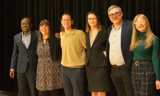 Six people with their arms around one another smiling on a stage in front of a black curtain. 