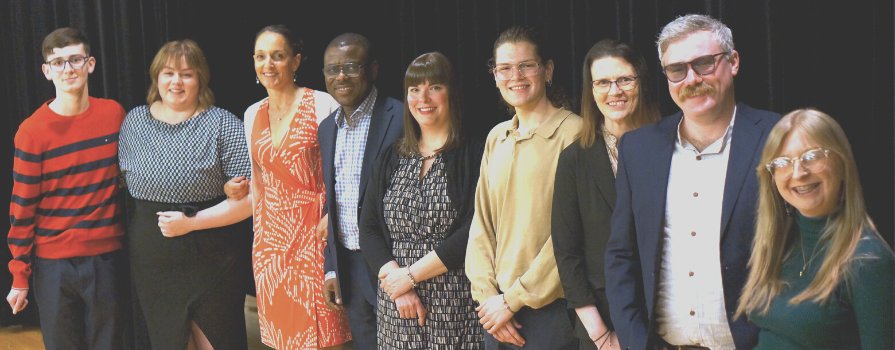 Nine people with their arms around one another smiling on a stage in front of a black curtain. 