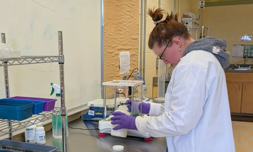 A women in a lab wearing a lab coat and purple gloves, working with laboratory equipment on a metal table.