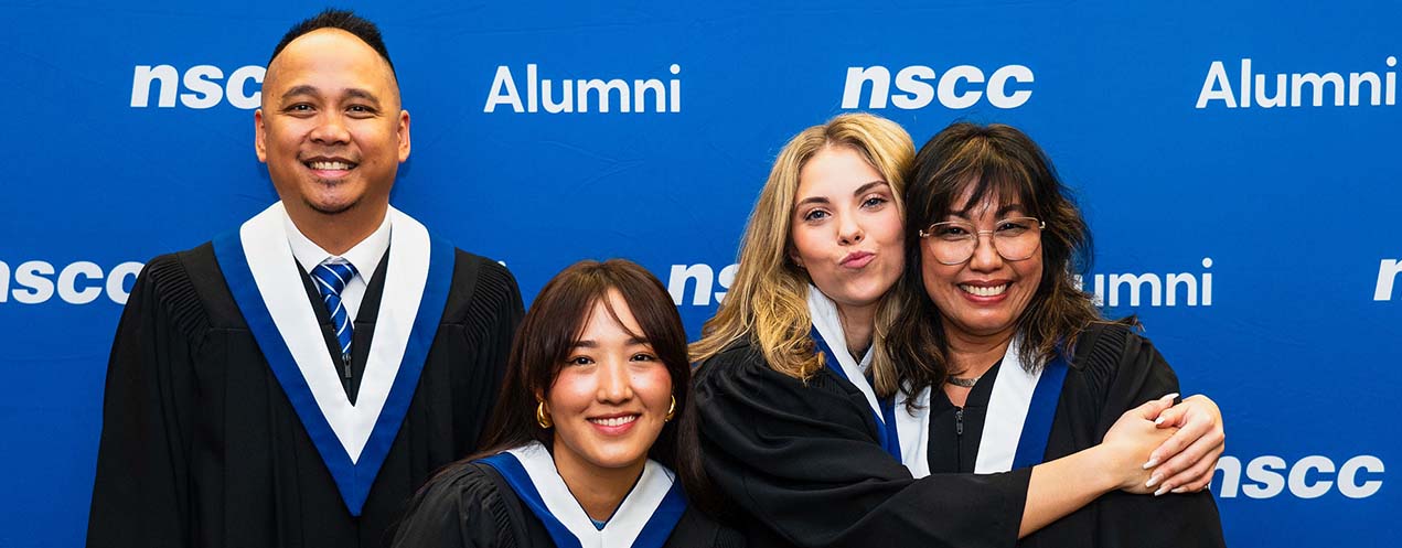 Four NSCC alumni stand, pose and smile in the Alumni photo area during Convocation. The banner behind them reads NSCC Alumni. They are wearing academic stoles and black gowns