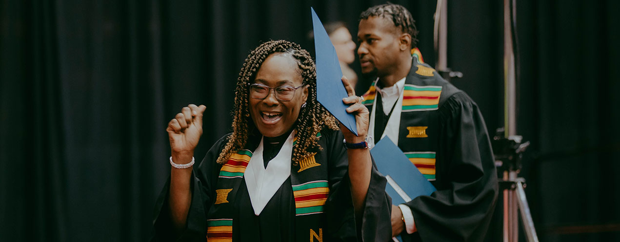 A woman wearing a convocation gown and Kente cloth sash celebrates with her new diploma at Convocation..