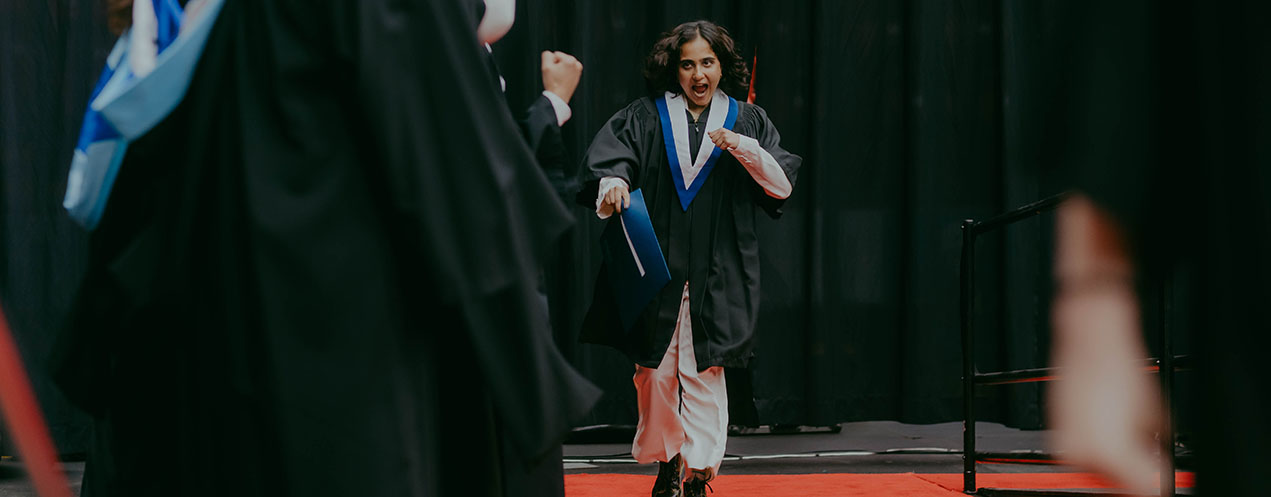 An NSCC graduate dances their way across the Convocation stage as they receive their credential. They are wearing their NSCC academic stole and graduation gown. They look very excited.