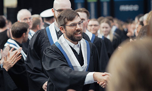 A smiling male graduate in glasses is shaking hands in his gown and stole.
