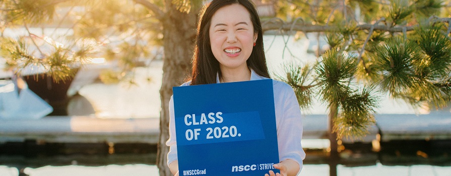 A woman holding a sign that says class of 2020 with a sunset and the ocean in the background.
