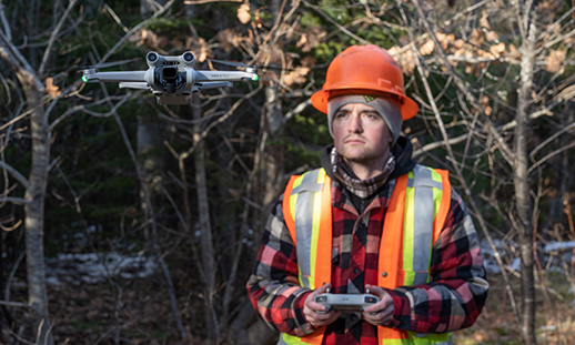 A man wearing forestry safety gear pilots a drone in the forest.