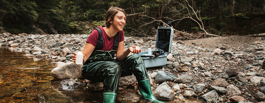 A woman smiles while examining a water sample from a river in the forest.