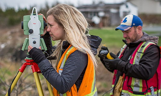 A woman uses a piece of surveying equipment while a man holds equipment in the background.