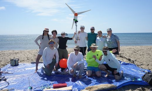 A group photo of 11 people, both women and men, standing and kneeling on a blue tarp on a beach in France with their floating structures for wind turbines.