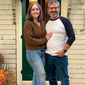 A woman and man standing together, smiling for a photo. They appear to be outside the front door of a house.