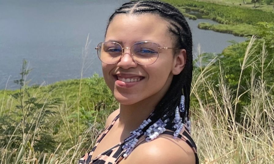 A female student smiling for a photo. She is outside in tall grass near a body of water.