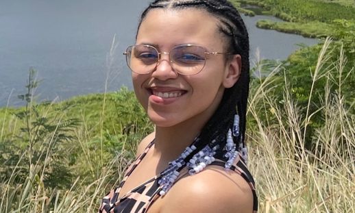 A female student smiling for a photo. She is outside in tall grass near a body of water.