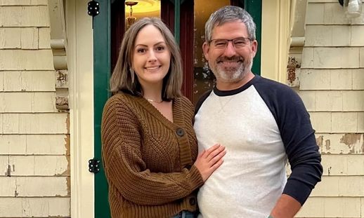 A woman and man standing together, smiling for a photo. They appear to be outside the front door of a house.