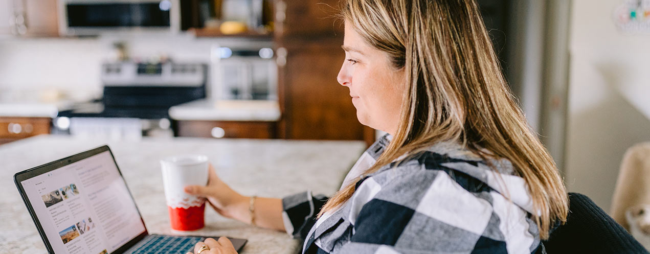 An eCampus learner sits in her kitchen in front of an open laptop while holding a cup in her right hand.