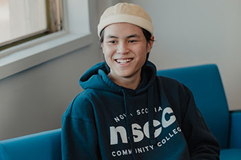 A student wearing a hat and a hoodie with the NSCC logo smiles while looking slightly to his right.