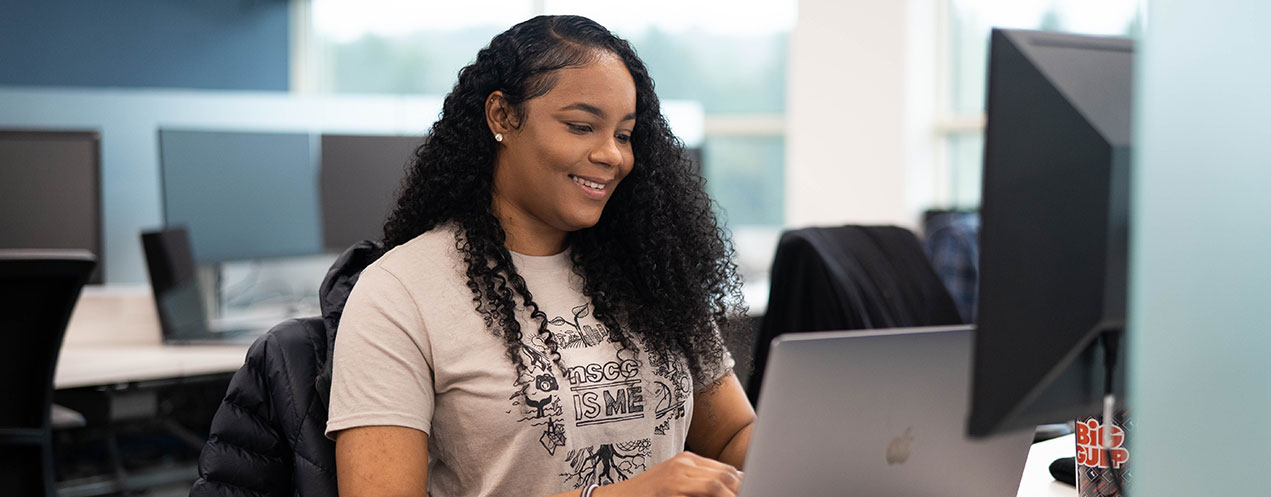 A student sits at a computer and types while smiling.
