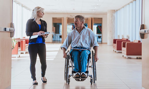 Woman walking beside a man in a wheel chair, approaching a wide open door in a hallway