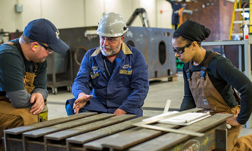 An instructor wearing a hard hat talking to a male and female student next to machine shop equipment