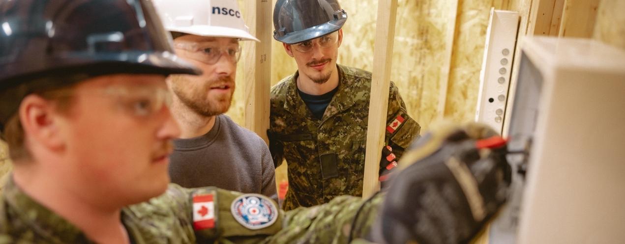 Faculty member and two military students inspect electrical work during hands-on training in a wooden shed.