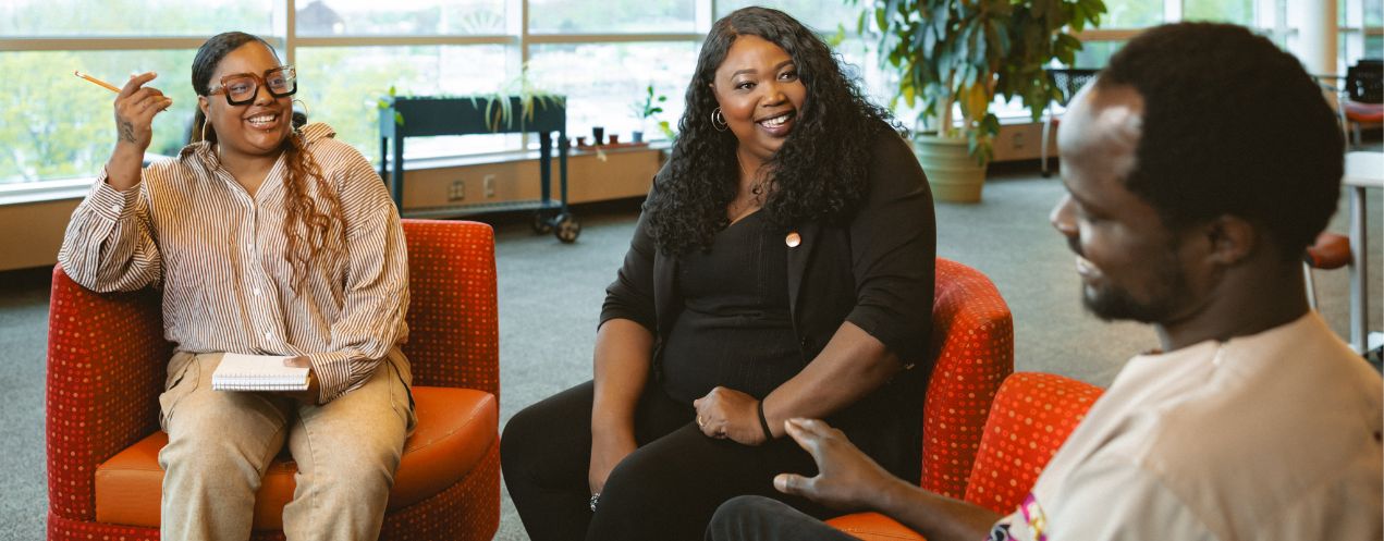 Three people are sitting together on chair in a bright library setting, with books and notepads with them.